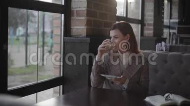 有魅力的女孩喜欢在咖啡休息时喝卡布奇诺，坐在一张靠窗户的桌子旁，坐在昂贵的桌子旁